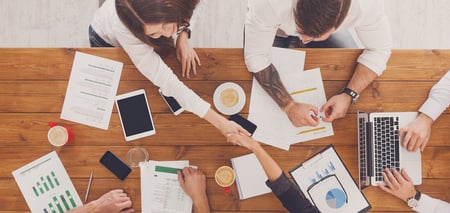Overhead view of four people working at a wooden table covered with financial statements, charts, a laptop and coffee, two of them shaking hands